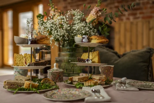 A tiered afternoon tea stand featuring a selection of savoury sandwiches including egg mayonnaise with rocket, cheddar with chutney, and ham with mustard mayonnaise, alongside sausage rolls and cheese rolls. The stand is served with a homemade scone topped with clotted cream and jam, and an assortment of assorted cakes, accompanied by tea and coffee in elegant crockery.