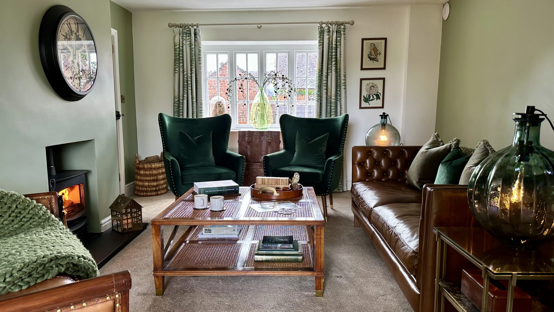 A cozy living room with sage green walls, a lit wood-burning stove on the left, and a large window letting in natural light. Two dark green velvet armchairs sit in front of the window, facing a wooden coffee table with books and mugs. A brown leather sofa with green accent pillows is on the right, next to a glass lamp on a side table. Botanical artwork hangs on the wall, and soft furnishings in green and brown tones create a warm, inviting atmosphere.