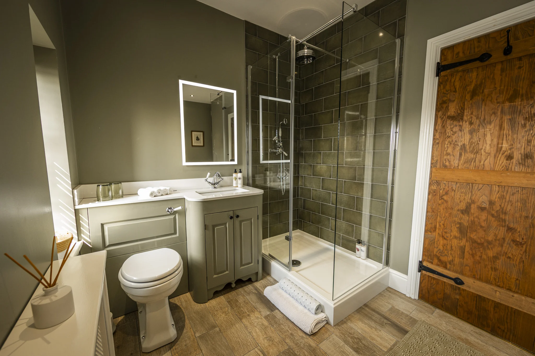 Stylish shower room at Owl Cottage, Peplow Hall Estate, Shropshire, featuring a walk-in glass shower, green tiled walls, vanity unit and rustic wooden door in a luxury self-catering holiday cottage.