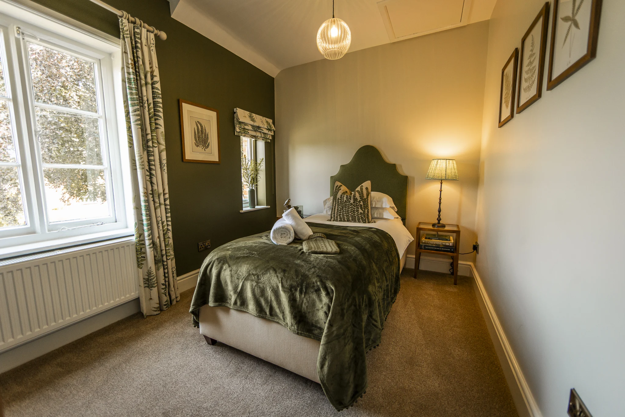 Elegant single bedroom at Owl Cottage, Peplow Hall Estate, Shropshire, featuring a green upholstered headboard, botanical decor, bedside lamp and large window in a luxury self-catering holiday cottage.