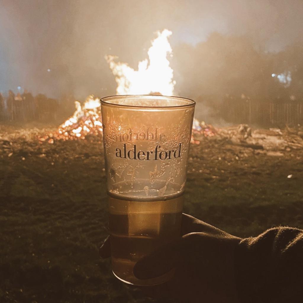 A hand holds a clear cup of beer labeled "alderford" in front of a large bonfire. The flames of the fire are burning brightly in the background, casting a warm glow and lighting up the smoky evening atmosphere. People can be seen standing around the bonfire in the distance, though they are blurred by the smoke and heat. The image captures the cozy and festive mood of an outdoor gathering, likely during a chilly night.