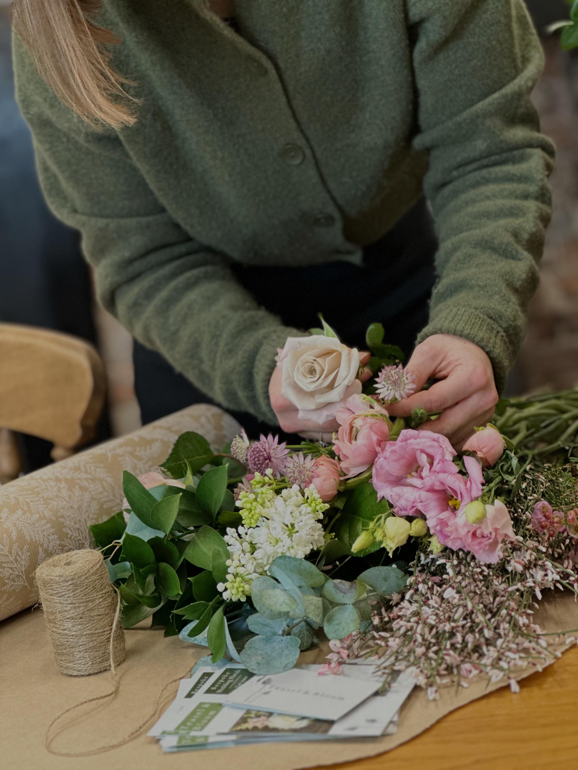 Person arranging a beautiful bouquet of fresh flowers, including roses, eucalyptus, and other seasonal blooms, on a rustic table with twine and wrapping paper nearby, creating a romantic and natural aesthetic