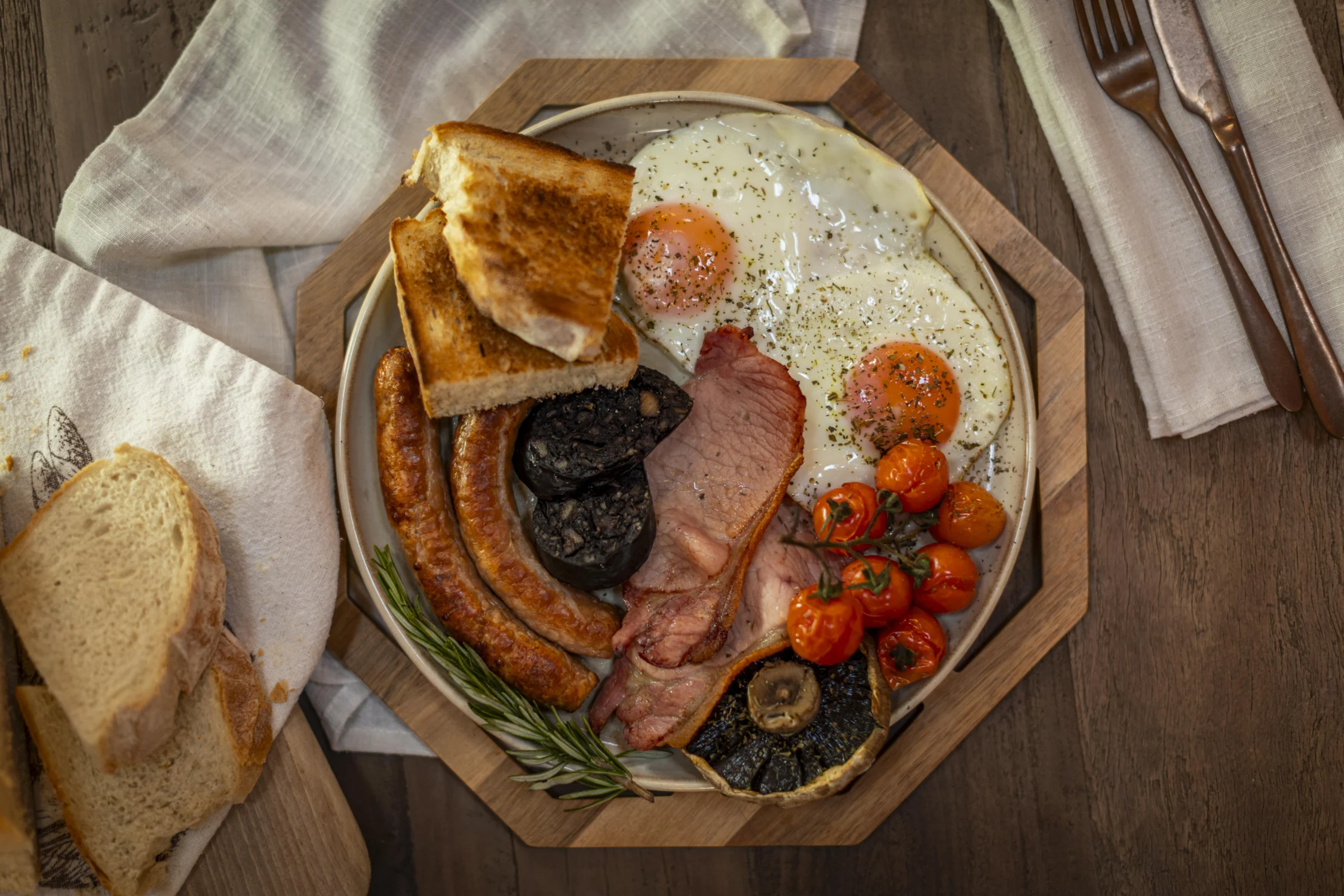 English Breakfast Full English breakfast on a ceramic plate: two sunny-side-up eggs seasoned with herbs, back bacon, two long sausages, grilled portobello mushrooms, black pudding, roasted cherry tomatoes on the vine, and buttered toast triangles. The plate sits on a wooden board with a sprig of rosemary, surrounded by a linen napkin, sliced bread, and copper cutlery on a dark wood table.