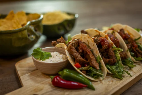 Three beef tacos in soft flour tortillas filled with seasoned steak strips, arugula, and chunky salsa. Served on a wooden board with a small bowl of sour cream garnished with cilantro, and fresh red and green chilli peppers. Bowls of tortilla chips are blurred in the background.