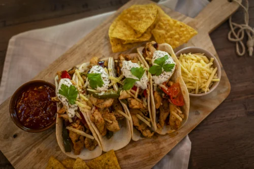 Four chicken fajita tacos in soft flour tortillas, filled with seasoned chicken, sautéed peppers, shredded cheese, sour cream, and fresh cilantro. Served on a wooden board with a bowl of salsa, a bowl of shredded cheddar, and scattered tortilla chips on a dark wood table.