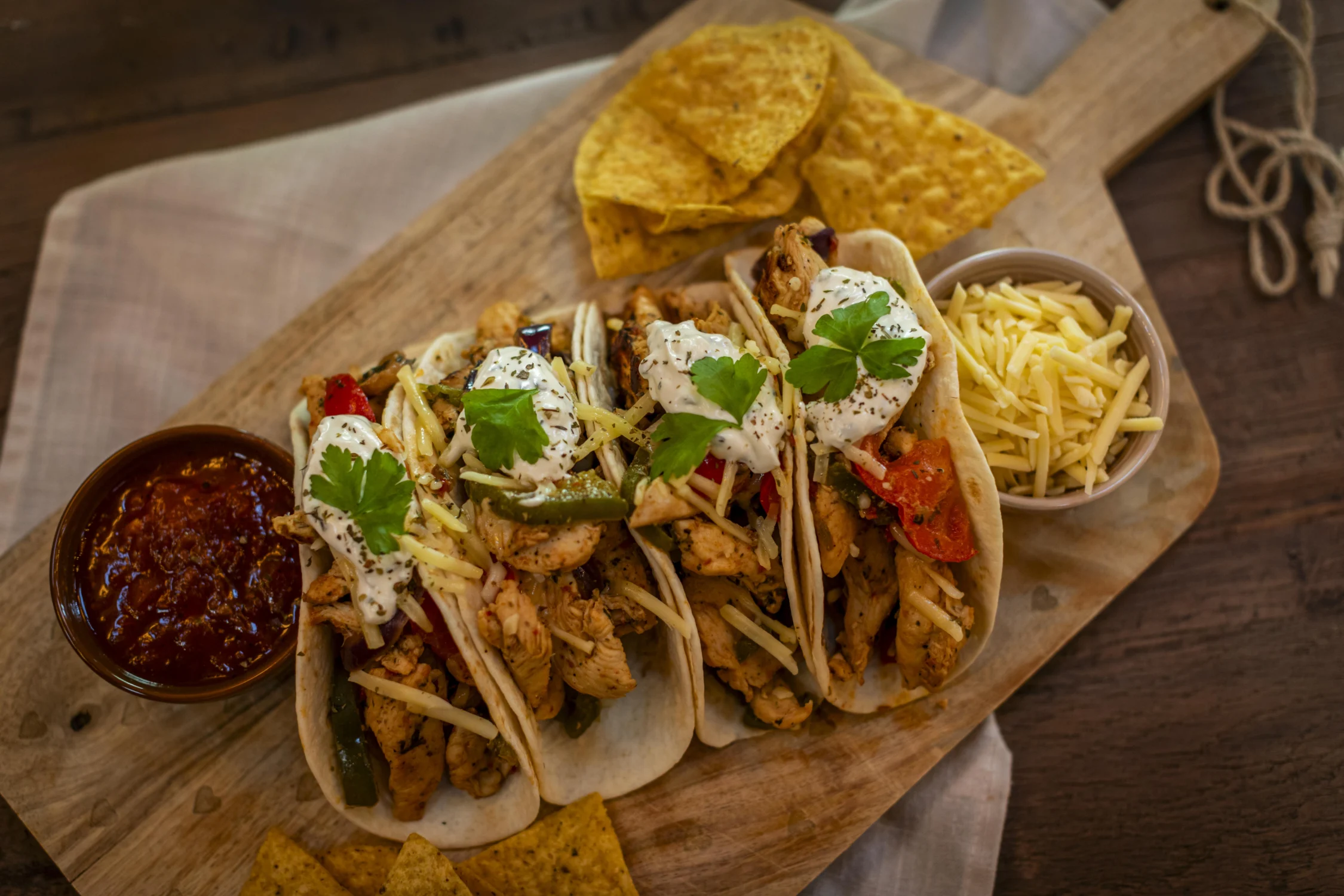 Chicken Fajitas Four chicken fajita tacos in soft flour tortillas, filled with seasoned chicken, sautéed peppers, shredded cheese, sour cream, and fresh cilantro. Served on a wooden board with a bowl of salsa, a bowl of shredded cheddar, and scattered tortilla chips on a dark wood table.
