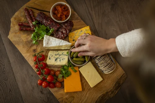 A hand reaches for a small bowl of pickles on a rustic wooden charcuterie board arranged with assorted cheeses, cured meats, crackers, cherry tomatoes, parsley, and a condiment, set on a dark wood table.