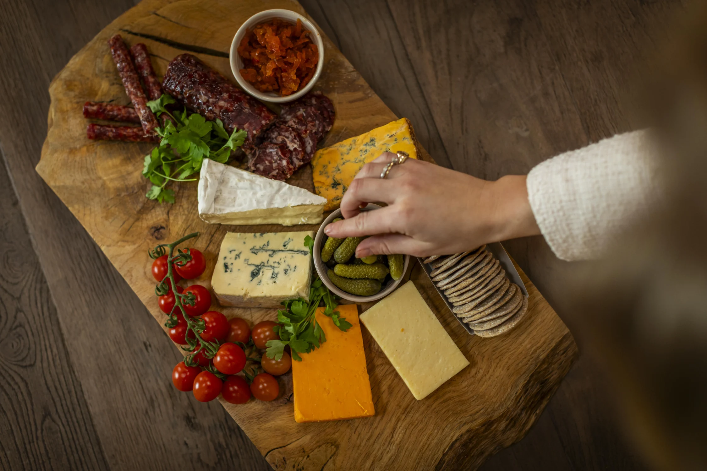 A hand reaches for a small bowl of pickles on a rustic wooden charcuterie board arranged with assorted cheeses, cured meats, crackers, cherry tomatoes, parsley, and a condiment, set on a dark wood table.
