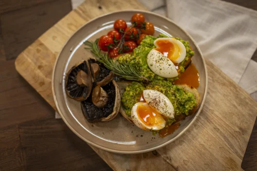 **Alt text:** Plate of avocado toast topped with soft-boiled eggs and herbs, served with roasted mushrooms, cherry tomatoes on the vine, and a sprig of rosemary on a beige ceramic plate atop a wooden board.