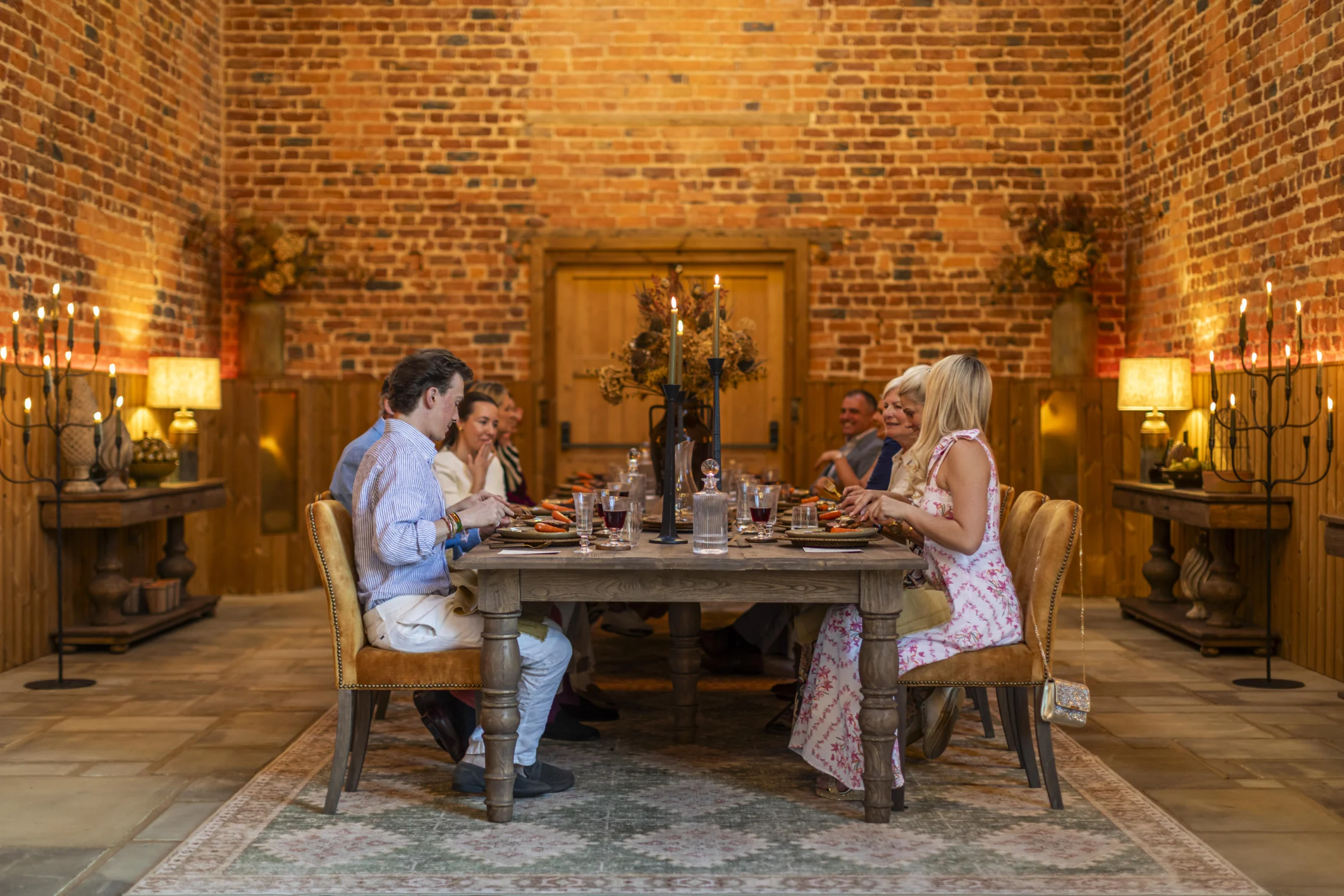 Fine Dining Table Guests seated around a long wooden dining table enjoy a meal in a warmly lit brick-walled barn. Tall candleholders, rustic furnishings, and soft ambient lighting create an elegant and intimate atmosphere, while diners converse and share food in a beautifully restored venue.