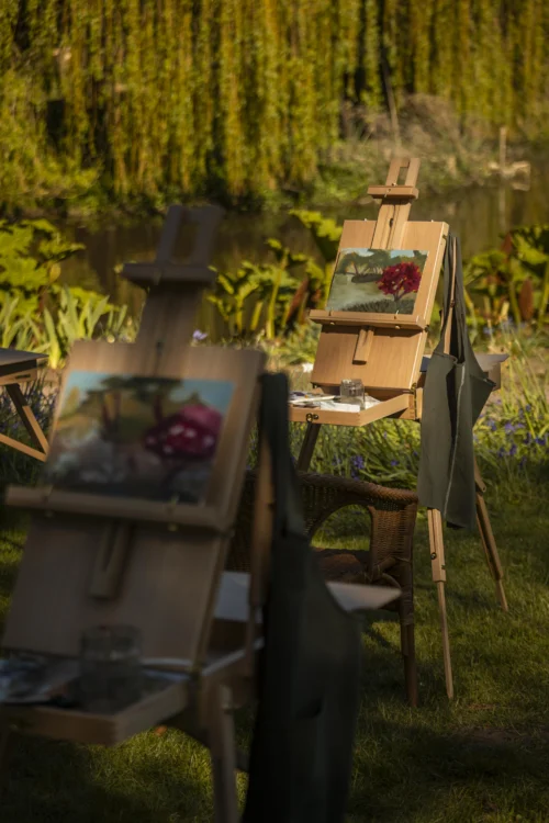 Several wooden easels are set up outdoors beside a lake, each displaying landscape paintings in progress. The scene is surrounded by lush greenery and hanging tree branches, with artist aprons draped over the easels. Sunlight filters through the garden, creating a peaceful atmosphere for an outdoor painting session.