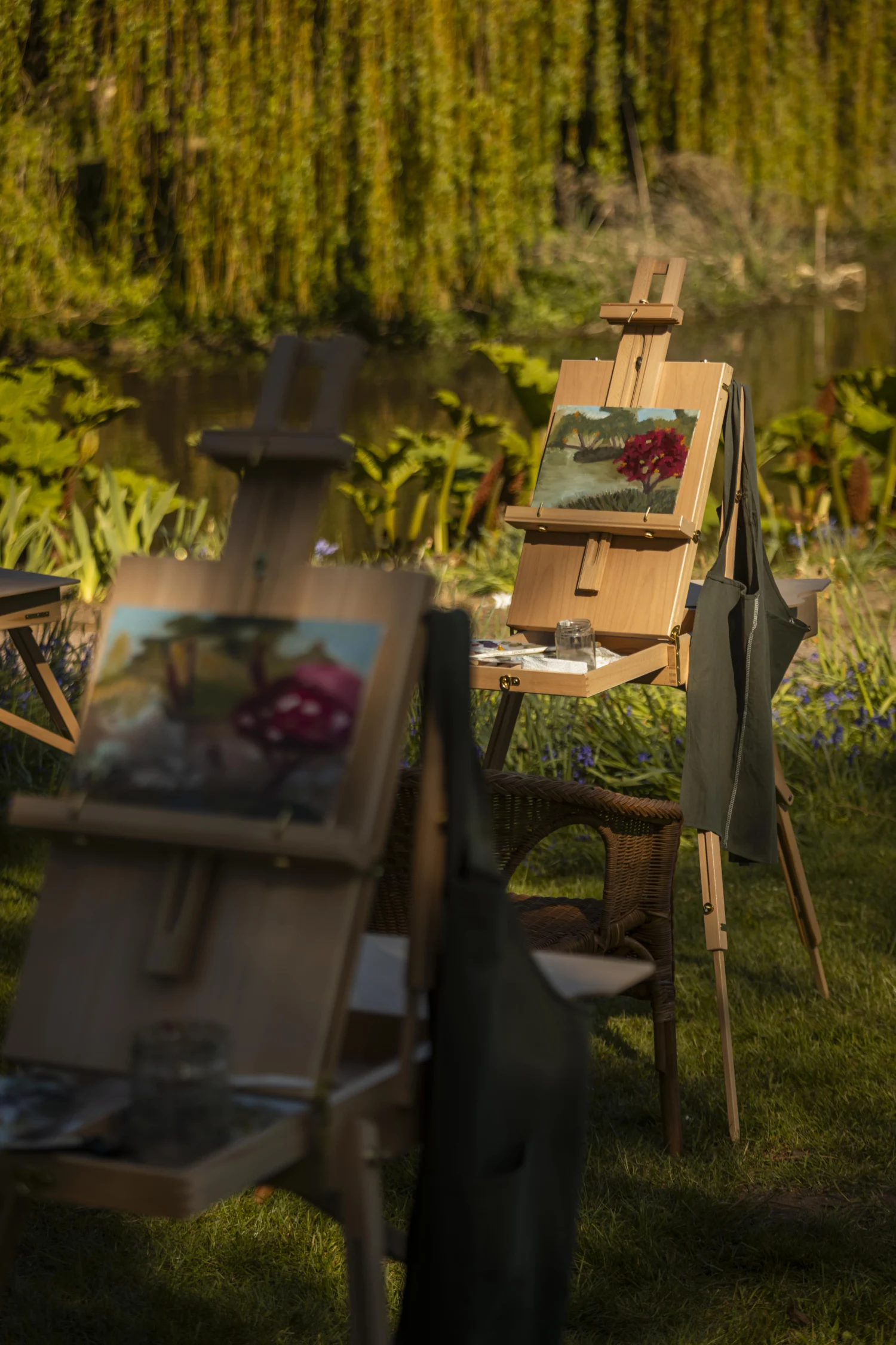 Oil Painting Several wooden easels are set up outdoors beside a lake, each displaying landscape paintings in progress. The scene is surrounded by lush greenery and hanging tree branches, with artist aprons draped over the easels. Sunlight filters through the garden, creating a peaceful atmosphere for an outdoor painting session.