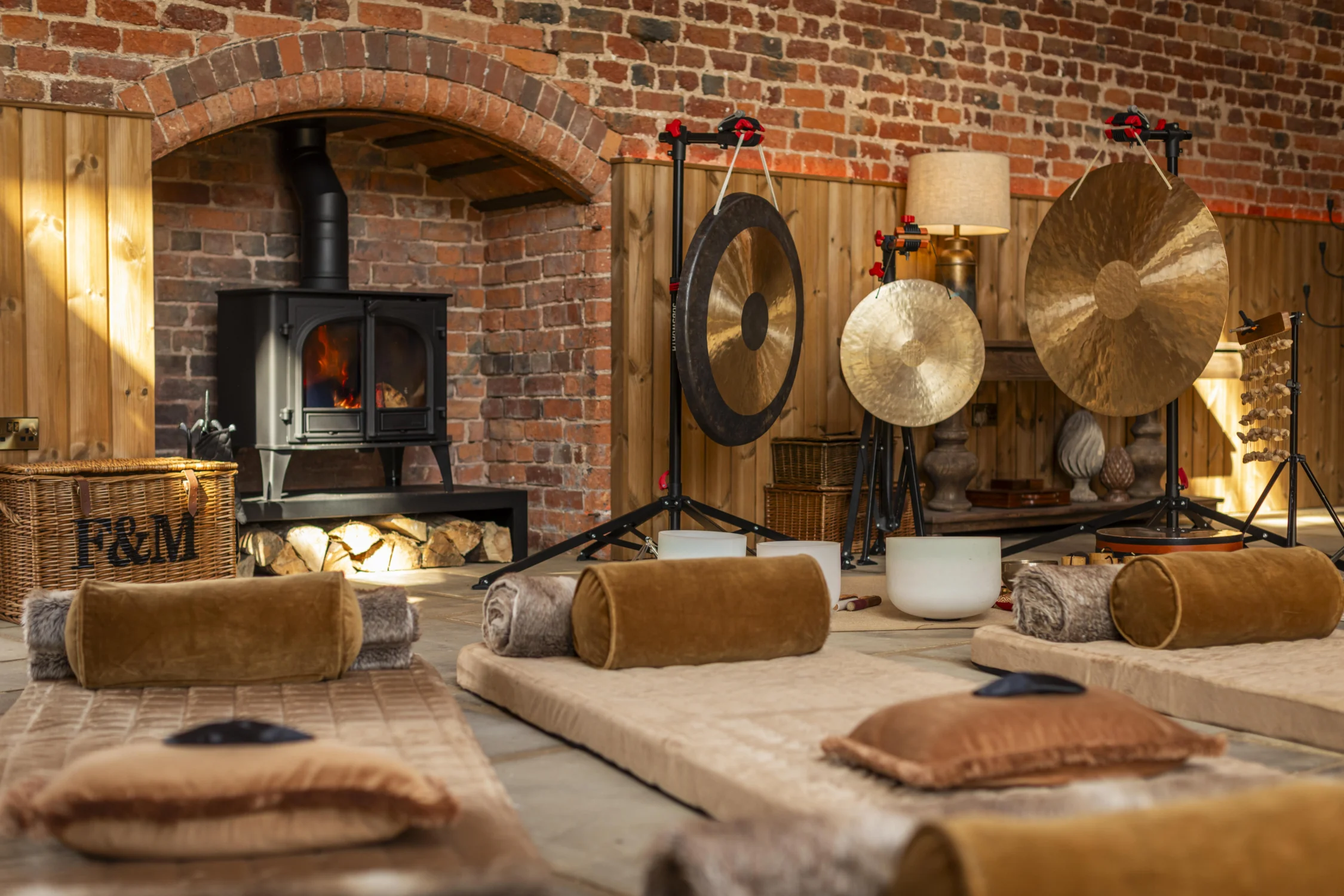 Sound bath setup with floor mats, gongs, crystal singing bowls, and a wood-burning stove in a warm, rustic room.