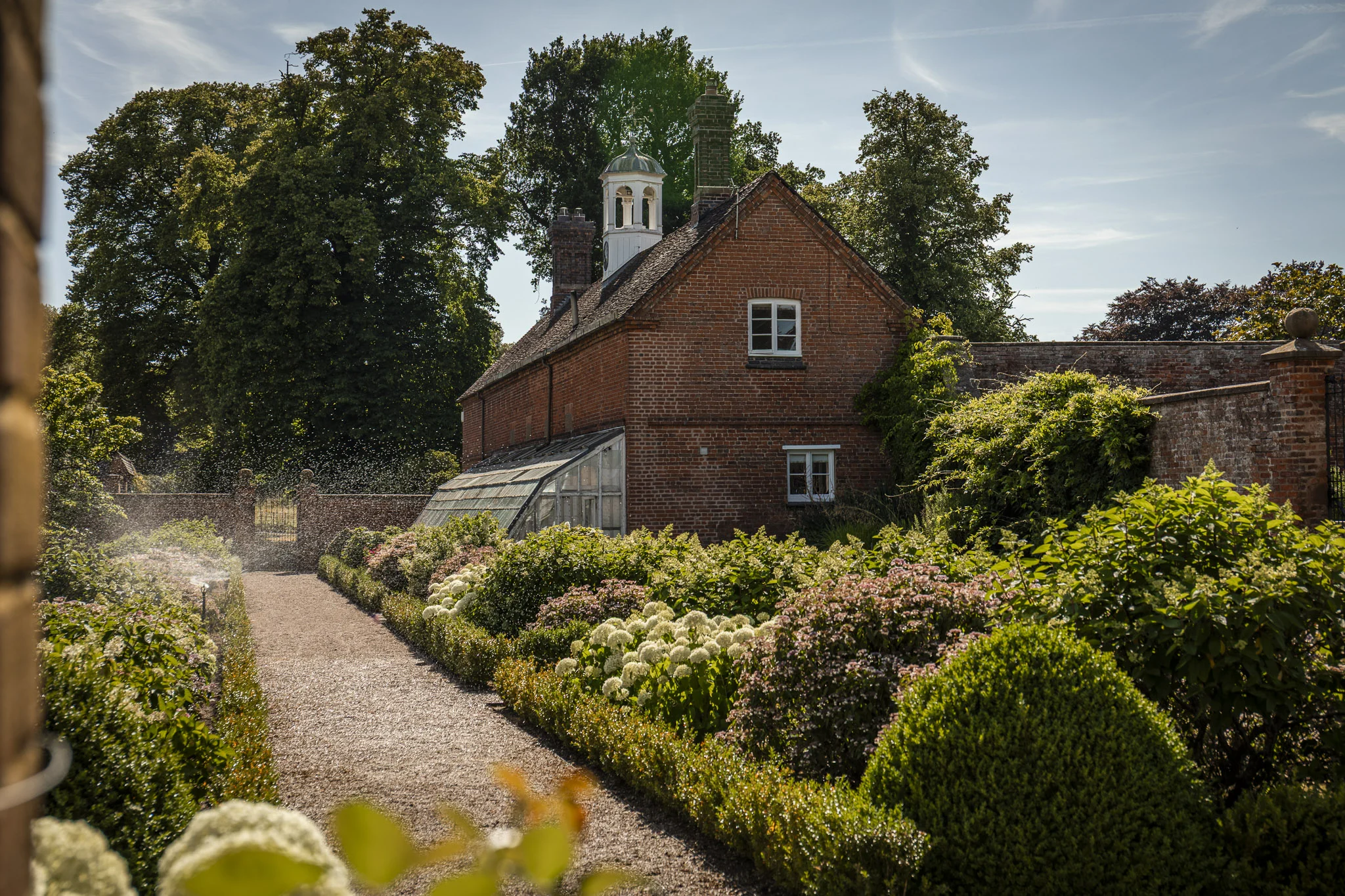 A red brick cottage with tall chimneys sits beside a lush, landscaped garden featuring neatly trimmed hedges, flowering shrubs, and a gravel path leading to a brick gate. A glass greenhouse is attached to the side of the house, while mature trees and a blue sky with wispy clouds create a peaceful countryside setting.
